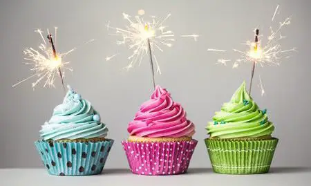 Three birthday cupcakes, each with a different color frosting: blue, pink and green. Instead of candles each has a burning sparkler placed at an angle on top.